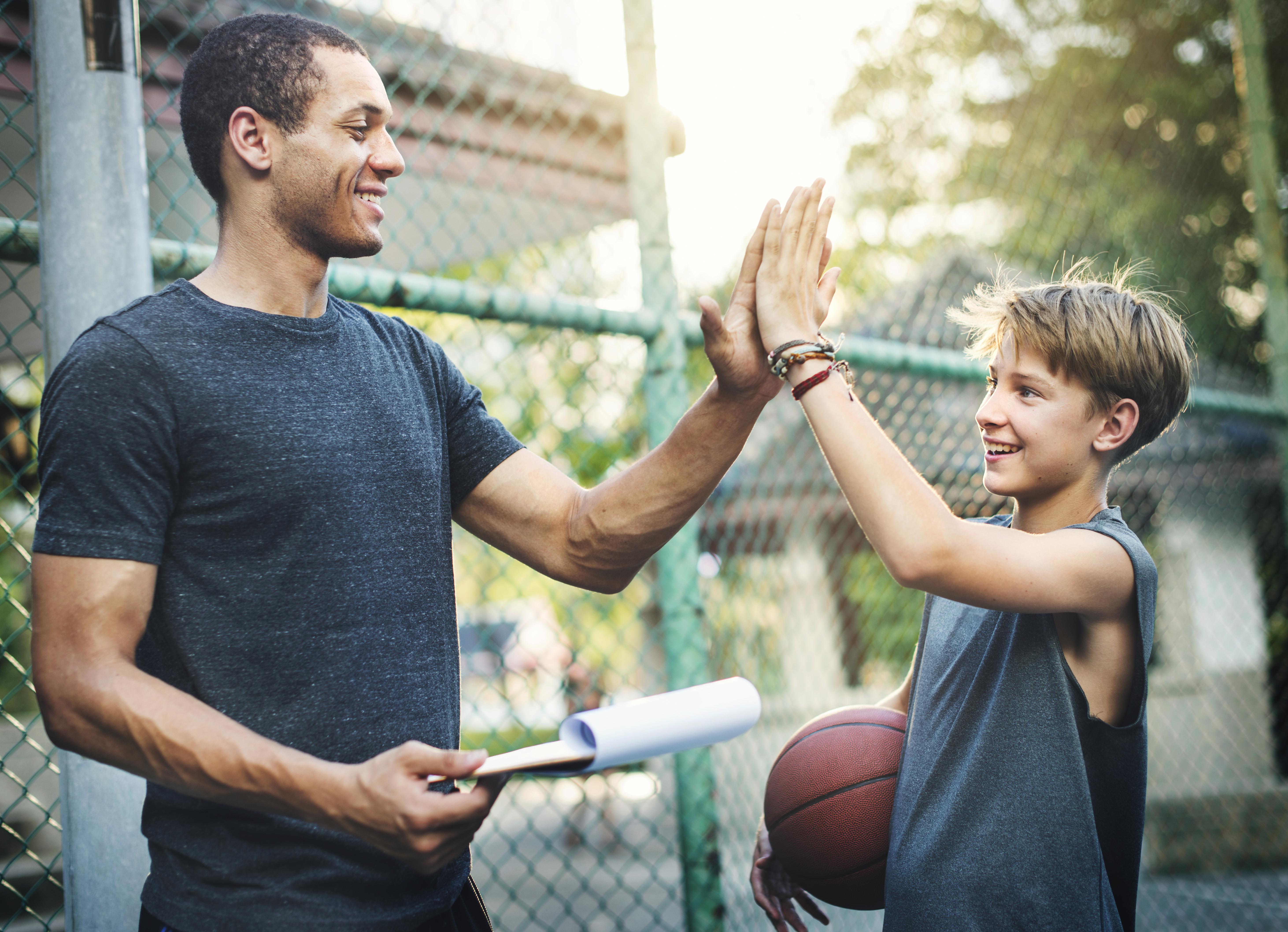 A young boy carrying a basketball is high-fiving an adult male carrying a clipboard on an outdoor basketball court.