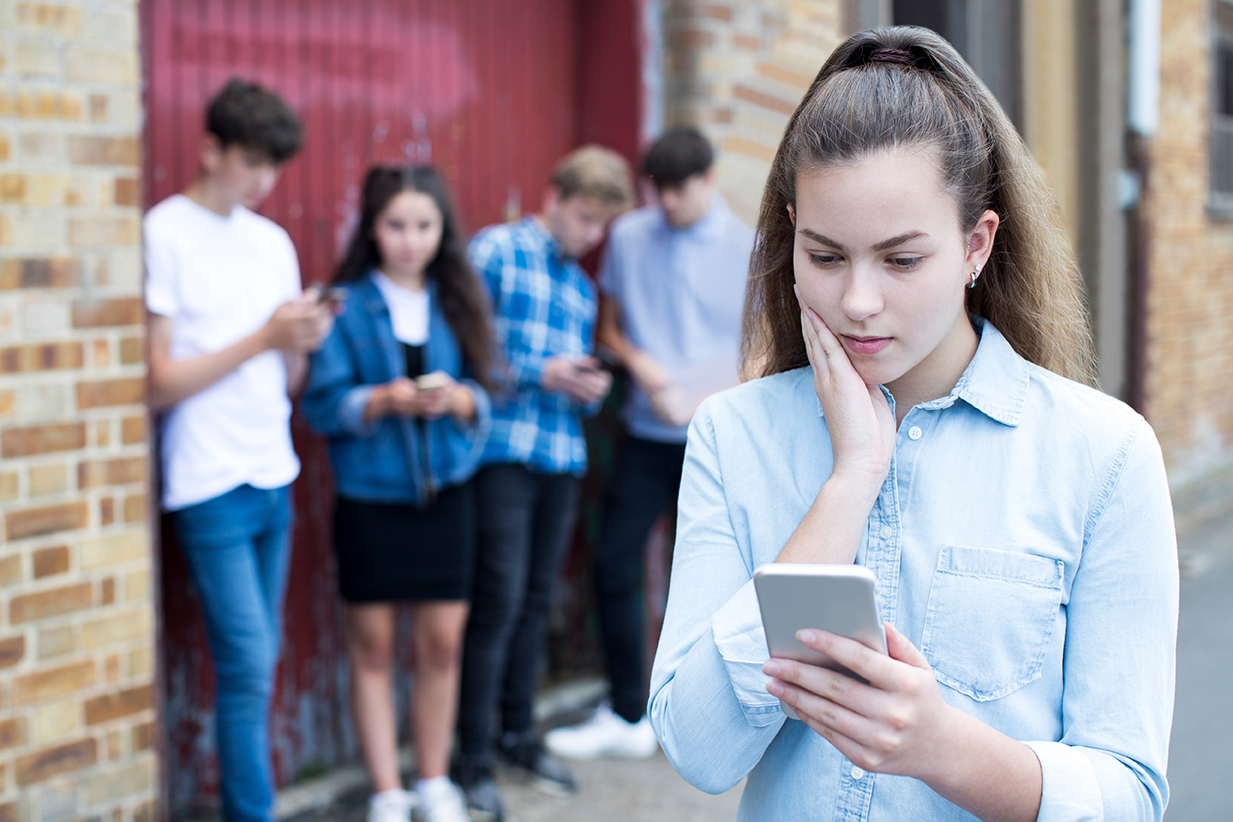 A teen girl looks at her cellphone with concern, as other teens gather and look at their cellphones in the background.