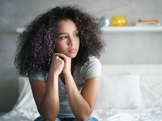 A teenage girl sits on the edge of a bed resting her head in her hands as she looks off. Her expression conveys worry, sadness, and thoughtfulness.
