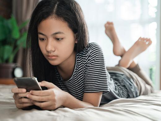 A teen girl plays on her phone while relaxing on a bed.