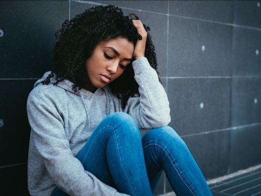 A teen girl looks sad as she sits on the ground against a wall and rests her head in her hand.
