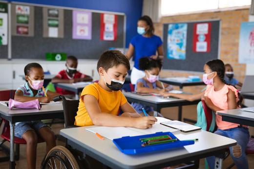 A child in a wheelchair sits at a desk in a classroom with other children and a teacher all wearing masks.